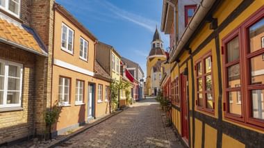 Geplaveide straat met kleurrijke vakwerkhuizen in geel, rood en bruin. Een kerktoren rijst op in de achtergrond onder blauwe lucht.