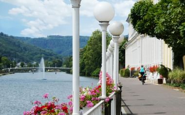 Elegante witte zuilengang met bollampjes langs de Lahn in Bad Ems. Kleurrijke bloemen langs de promenade met zichtbare fietsers.
