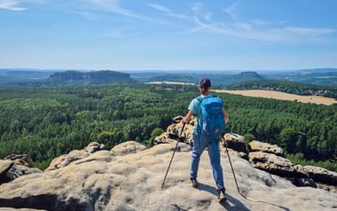 Wandelaar met blauwe rugzak en wandelstokken op rotsformatie bij Gohrisch, uitzicht over boslandschap met velden.