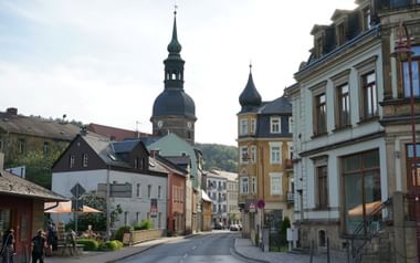 Straatbeeld in Bad Schandau met kleurrijke historische gebouwen en kerk met donkere koepeltoren. Voetgangers lopen langs de gebogen weg.