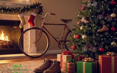 Versierde kerstboom met fietsversieringen naast een vintage bruine fiets. Ingepakte cadeaus en wandelschoenen voor een open haard.