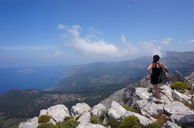 Wandelaar op rotsachtige bergtop op Rhodos met uitzicht op blauwe Egeïsche Zee, groene valleien en verre kustlijn onder heldere hemel.