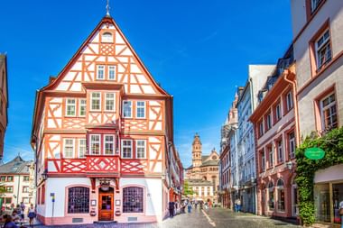 Geplaveide straat in de oude binnenstad van Mainz met kleurrijke vakwerkhuizen en barokke domtorens zichtbaar op de achtergrond.