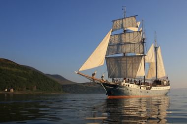 Driemast zeilschip Atlantis met witte zeilen op kalm water bij groene heuvels onder blauwe hemel bij zonsondergang.