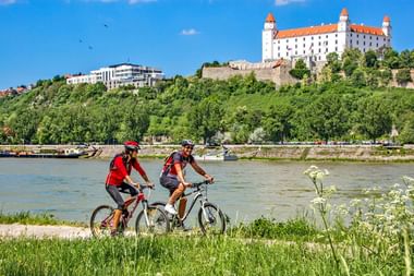Twee fietsers in rode shirts met fietsen op grasrijke Donauoever. Kasteel van Bratislava met witte muren en rood dak zichtbaar op heuvel.