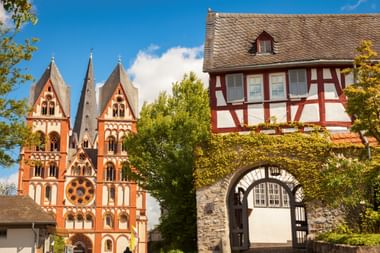 Rood-witte Dom van Limburg met drie torens naast een traditioneel vakwerkhuis met stenen boog, omringd door groene bomen onder blauwe lucht.
