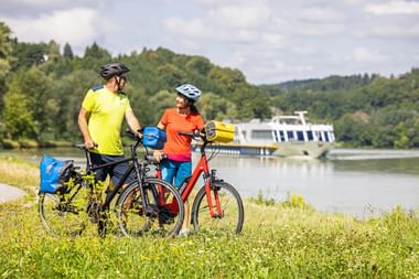 Twee fietsers met helmen nemen een pauze bij de Donau met hun toerfietsen. Een passagiersschip is zichtbaar op het water.