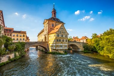 Historisch vakwerkhuis van het Oude Stadhuis van Bamberg op eiland in de Regnitz, verbonden door stenen brugbogen onder blauwe lucht.