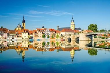 Historische stenen brug over de Main in Kitzingen met kleurrijke gebouwen en kerktorens weerspiegeld in kalm water onder blauwe lucht.
