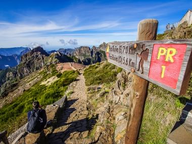 Houten wegwijzer op Madeira toont PR1 wandelroute met afstanden naar Pico Ruivo. Wandelaar zit ernaast met uitzicht op dramatische bergtoppen.