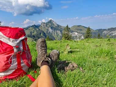 Bruine wandelschoenen en rode rugzak op groen gras met Kampenwand bergtop en alpenlandschap op de achtergrond.