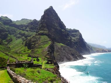 Kaapverdisch kustpad met dramatische kliffen Stenen wandelpad langs de kust van Kaapverdië met hoge vulkanische kliffen, turquoise oceaan en oude stenen ruïnes op groene heuvel.
