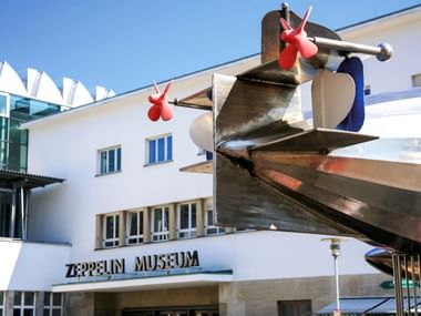 Wit modernistisch gebouw met Zeppelin Museum bordje en grote metalen propellersculptuur met rode accenten boven de ingang.