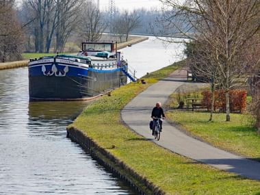 Fietser rijdt over verharde pad langs kanaal in Elzas met afgemeerde vrachtschepen en kale winterbomen langs de waterweg.