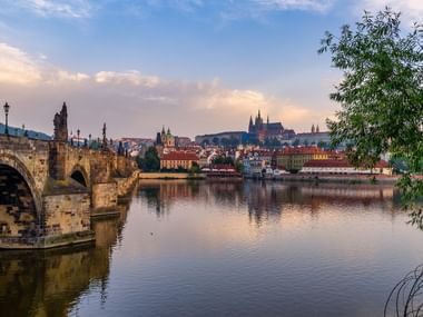 De historische Karelsbrug overspant de Moldau in Praag met de Praagse Burcht zichtbaar op de heuvel. Avondlicht weerkaatst op kalm water.
