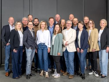 Groep van ongeveer 20 professionals in business casual kleding die samen staan voor een grijze muur in een moderne kantooromgeving.