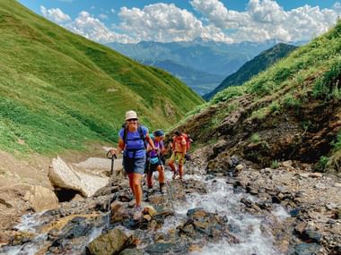 Groep wandelaars met rugzakken en wandelstokken steekt rotsachtige beekbedding over in Georgische bergen met groene heuvels.