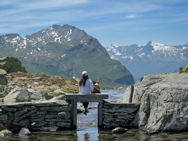 Vrouw in wit shirt zit met hond op stenen brug over bergbeek bij Rusettameer. Besneeuwde toppen van Via Spluga zichtbaar op achtergrond.