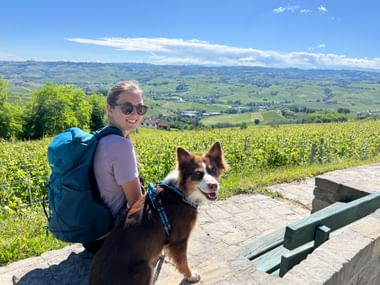Isabella met rugzak zit naast haar hond Leyla op stenen uitkijkpunt met uitzicht op Piëmontese wijnlandschap met groene heuvels.
