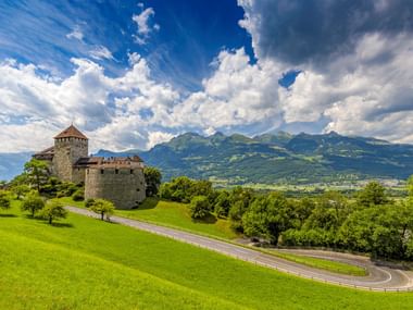 Kasteel Vaduz met Alpenlandschap Kasteel Vaduz op een groene heuvel in Liechtenstein met kronkelende weg, omgeven door weelderige weiden en Alpengebergte.