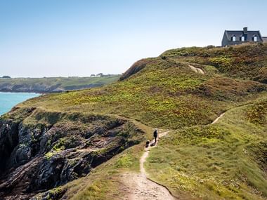 Persoon wandelt met hond op kronkelend kustpad langs groene kliffen met uitzicht op turkooizen zee, huis zichtbaar op heuvel.