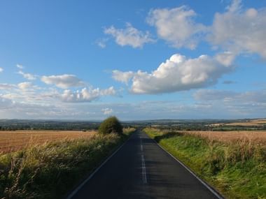 Landweg door het Cotswolds platteland Rechte landweg in de Cotswolds met gouden velden aan beide kanten, groene hagen en blauwe lucht met witte wolken.