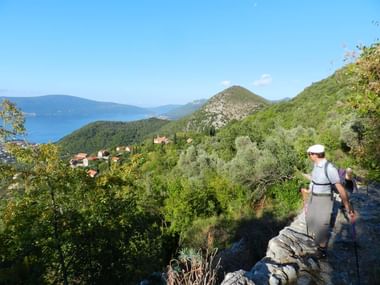 Wandelaars op rotsachtig kustpad in Montenegro met groene heuvels, dorpshuizen en blauwe zee op de achtergrond onder heldere hemel.