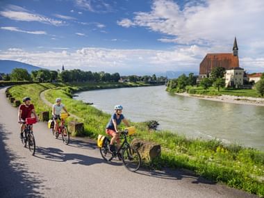 Drie fietsers rijden langs de Salzach bij Laufen. Een historische kerk met rood dak en toren staat aan de overkant van de rivier.