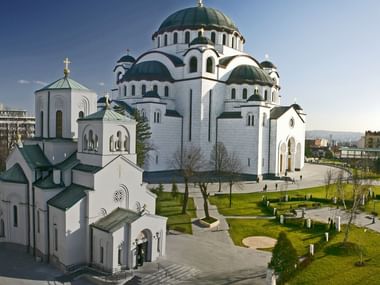Grote witte orthodoxe kathedraal met groene koepels en gouden kruisen in Belgrado, omgeven door aangelegde tuinen en stedelijke gebouwen.