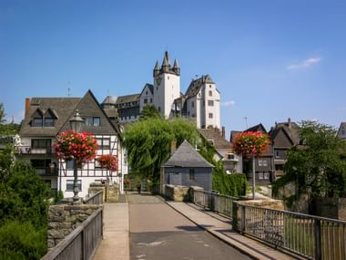 Historische brug naar kasteel Diez met witte torens. Traditionele vakwerkhuizen met rode bloembakken langs de rivieroever onder blauwe lucht.