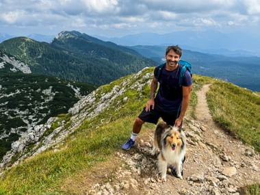 Glimlachende wandelaar met rugzak en Collie-hond op bergkampad in de Julische Alpen. Groene hellingen en bergtoppen onder bewolkte hemel.