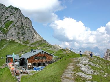 Alpenhut met houten gevel in groene weiden op de Tirolerweg, omgeven door dramatische kalkstenen pieken en wandelpaden.