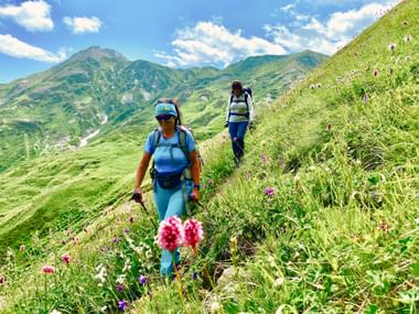 Twee vrouwelijke wandelaars met rugzakken lopen op een grasrijke berghelling in Georgië met wilde bloemen en groene bergketens onder blauwe lucht.