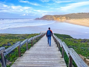 Wandelaar met rugzak op houten loopbrug naar strand op Rota Vicentina. Groene vegetatie langs het pad, oceaangolven en kliffen zichtbaar.