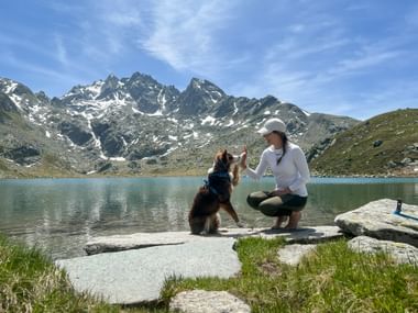 Vrouw in wit shirt en pet gehurkt met hond op rotsachtige oever van Rusettameer. Besneeuwde bergen en blauwe lucht op achtergrond.