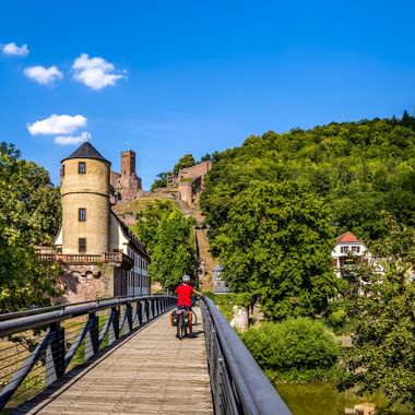 Fietser in rode jas steekt houten brug over in Wertheim. Historische toren en kasteelruïne op groene heuvel onder blauwe lucht.
