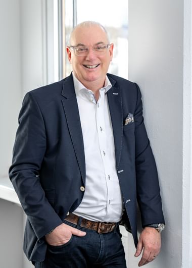 Edwin Schollen glimlachend, met bril, wit overhemd en marineblauw jasje met pochet, staand in licht kantoor met witte muren en raam.