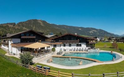 Hotel Quehenberger in Maishofen met groot buitenzwembad omgeven door groen gazon en houten hek, tegen Alpengebergte onder blauwe lucht.