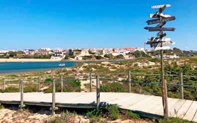 Houten richtingbord op kustloopbrug met uitzicht op baai met witte huizen en boten. Borden wijzen naar verschillende bestemmingen langs wandelpad.
