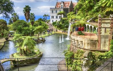 Terrasvormige tropische tuin in Funchal, Madeira met vijvers, fonteinen, weelderige vegetatie en wit paleis met rood dak.
