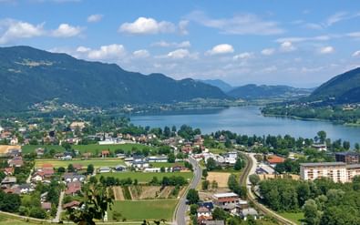 Panoramisch uitzicht op Ossiacher See vanaf Peterlewand met het meer omgeven door groene bergen, dorpen en wegen onder blauwe hemel.