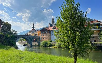 Stenen brug over rivier in Murnau met kleurrijke historische gebouwen, kerktoren en groene boom op voorgrond onder blauwe hemel.