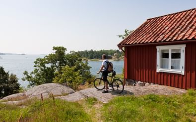 Fietser met fiets staand op rotsformatie naast traditioneel rood houten huisje met uitzicht op wateren van Stockholms archipel.