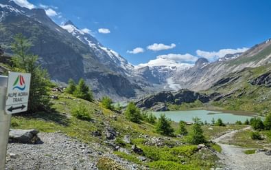 Alpe Adria Trail bord bij Margaritzensee meer met Grossglockner gletsjer en besneeuwde toppen onder blauwe lucht.