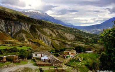 Klein bergdorp in Hidden Valley, Albanië met stenen huizen en terrasvormige velden, omgeven door geërodeerde hellingen en besneeuwde toppen.
