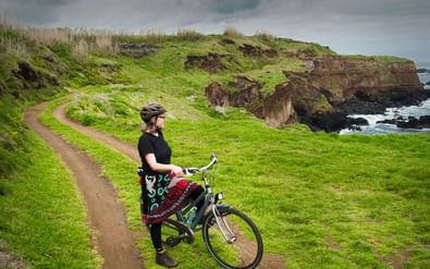Vrouwelijke fietser met helm staat naast fiets op grasachtig kustpad in São Miguel, Azoren, met dramatische kliffen en oceaan op achtergrond.