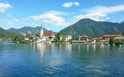 Schilderachtig uitzicht op Rottach-Egern aan het Tegernsee met witte kerktoren, traditionele gebouwen en groene bergen onder blauwe lucht.