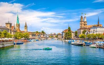 Limmat rivier door Zürich met boten aan de oevers, historische kerktorens en Grossmünster kathedraal onder blauwe hemel.