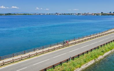 Luchtfoto van fietsers op een brug over een turquoise lagune bij Grado, Friuli. De kuststad is zichtbaar in de verte onder blauwe lucht.