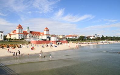 Kurhaus Binz spa hotel met kenmerkende rood-bedekte torens met uitzicht op een zandstrand aan de Oostzee met bezoekers en strandkorfjes op Rügen.